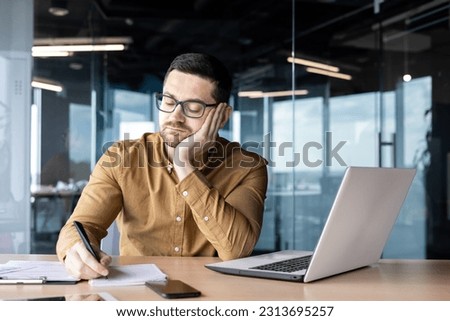 Bored and sad young man working in the office at a laptop and making notes in a notebook with a pen. Overworked, tired of work. Royalty-Free Stock Photo #2313695257