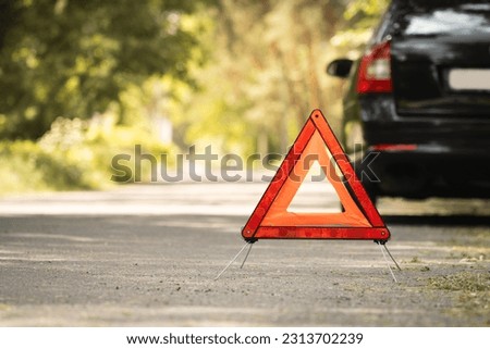 Red triangle, red emergency stop sign and black car with technical problems in the blurred background. Emergency stop of the car on the road. Safety procedure while having a vehicle broken down