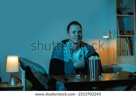 Man watching TV and eating popcorn at home at night. Portrait of a man sitting on a couch in a room filled with a blue screen light from a huge TV