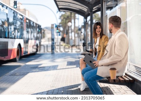 Two cheerful friends young woman and man sitting at bus station while city bus is passing by on the street, having a conversation, laughing, holding mobile phone and digital tablet. Copy space photo. Royalty-Free Stock Photo #2313705065