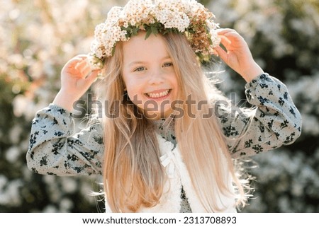 Smiling child girl wear floral wreath and dress posing over blooming tree outdoor at nature background. Looking at camera. Childhood. Springtime.  Royalty-Free Stock Photo #2313708893