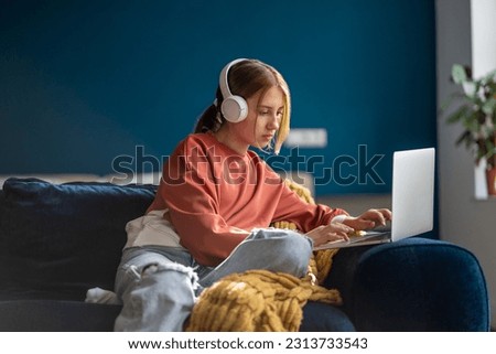 Focused teenage girl wearing headphones typing text on laptop relaxed sitting on couch in living room. Pensive teenager school student studying online, distance learning, using computer at home.