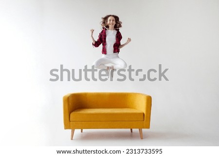 young cute girl jumps and rejoices in victory on soft comfortable sofa, woman celebrates success and good luck on yellow couch on white isolated background