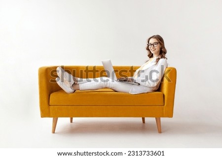 young businesswoman in suit uses laptop on comfortable soft sofa, girl in formal wear is typing on computer on yellow couch on white isolated background Royalty-Free Stock Photo #2313733601
