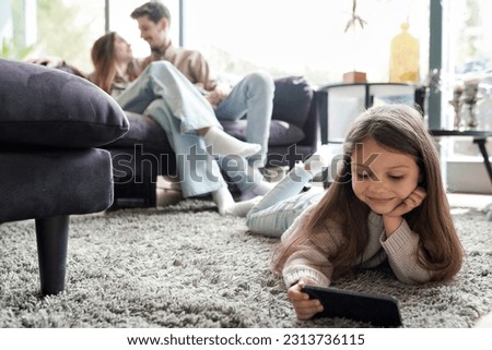 Portrait of little girl child watching video using mobile phone and wireless internet connection while lying on home floor. Young loving parents sitting on sofa couch hugging spending time together