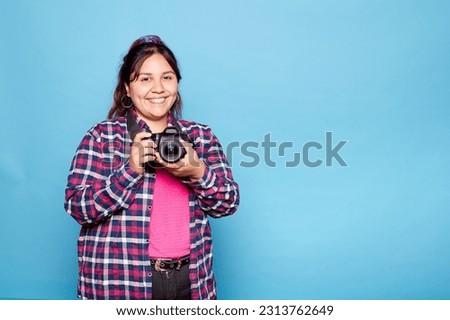 Young curvy latina woman smiling happy holding reflex camera isolated on blue background. Indoor studio shot. Copy space. Royalty-Free Stock Photo #2313762649