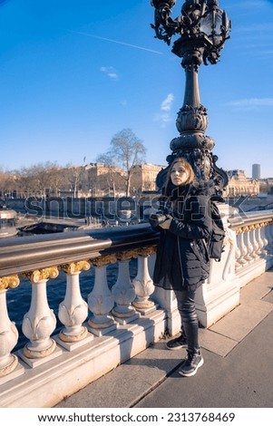 Full body of thoughtful and pensive young Latin woman traveler in warm outfit standing on Pont Alexandre bridge with photo camera and looking away during sightseeing trip in Paris, France