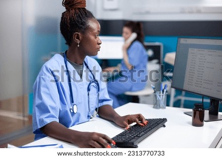 African american nurse working at desk, inserting data in appointment table list. Concentrated adult woman checking patient list, wearing stethoscope in clean, modern medical office operating computer Royalty-Free Stock Photo #2313770733