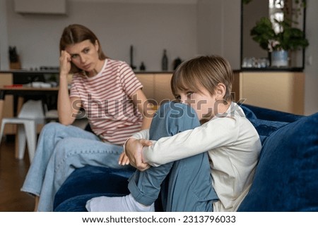 Frustrated teenage boy sits hugging knees on couch and looks away after argument with mother. Parent sits next to son and comforts after quarrel. Calming down child due to school problems Royalty-Free Stock Photo #2313796033