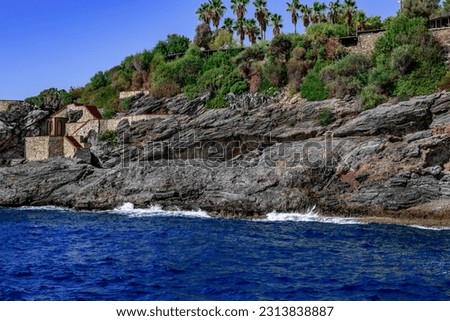 Layered granite cliff over the sea in the abandoned Emirgan Ulas Mesire Yeri Park in Alanya (Turkey). Stone steps in the rock against the background of blue water and tropical plants