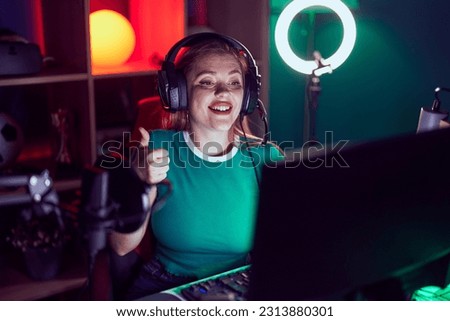 Redhead woman playing video games smiling happy and positive, thumb up doing excellent and approval sign 