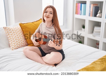 Young redhead woman playing ukulele sitting on bed at bedroom