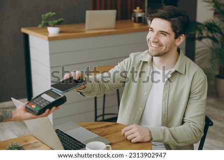 Young man wearing casual clothes sit alone at table in coffee shop cafe restaurant indoors work on laptop pc computer hold bank terminal process acquire mobile cell phone payment. Freelance concept Royalty-Free Stock Photo #2313902947