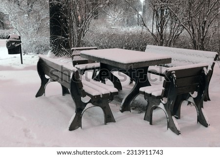 Black and white photo. Snow-covered benches and a table in the park in the evening
