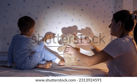 Smiling boy with mother showing theater of shadows with flashlight on the wall. Family having time together, parenting, happy childhood and entertainment