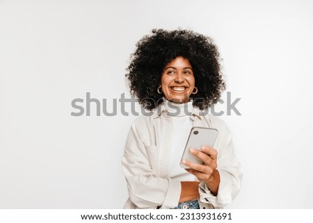 Happy young woman looking away with a smile while holding a smartphone in her hand. Beautiful woman with curly hair contemplating an idea while standing against a studio background.