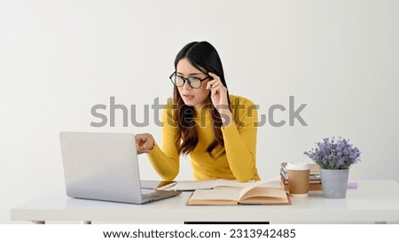 A puzzled and confused young Asian female college student is touching her glasses, looking at her laptop screen with hesitation, and sitting at her desk. isolated on a white background