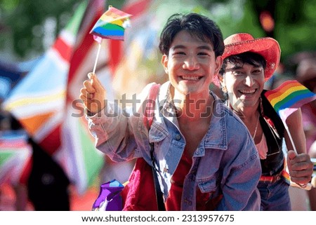 Gay couple waving rainbow flags in front of camera, as the Pride parade was moving down the streets in the background, celebrate LGBTQ Pride month parade, smiling and looking at camera