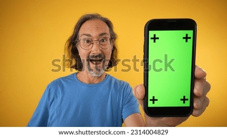 Excited mature elderly man standing and pointing Big smartphone with blank green screen with tracking points, demonstrating copy space for app or website design, mockup image on yellow background