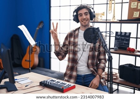 Young hispanic man playing piano at music studio smiling with happy face winking at the camera doing victory sign. number two. 