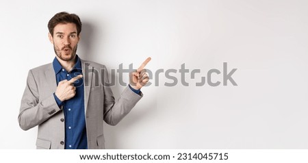 Excited handsome caucasian businessman in suit pointing right at logo, say wow and look amazed, standing on white background.
