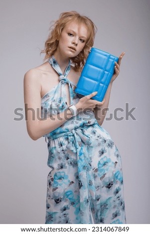 High fashion photo of beautiful elegant young woman in a pretty rich blue jumpsuit with floral pattern, handbag, clutch posing over white, soft gray background. Studio Shot, portrait. Blonde