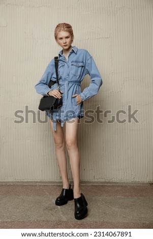 High fashion photo of a beautiful elegant young woman in pretty blue denim, jeans jumpsuit, black handbag. Sandy beige textured wall. Blonde, slim figure, short haircut