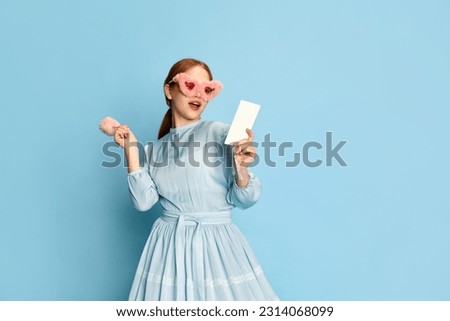 Portrait with young girl, princess with red hair wearing dress and pink plush sunglasses posing with an interested face over blue background. Concept of medieval era, beauty, modern fashion, ad