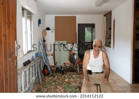 older man sitting on a stool posing with his onion harvest in a garage, person in his eighties wearing a white tank top and mobile phone holstered to his belt. Royalty-Free Stock Photo #2314112123