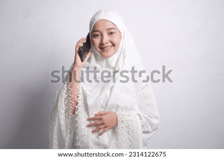 Smiling Asian Muslim woman wearing white mukena  currently calling  on mobile phone and looking empty space,eid al adha concept, isolated on white studio background