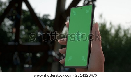 CU Silhouette of Caucasian female holding a generic smart phone in a right hand in vertical orientation, screen facing camera. Garden with green plants in the background