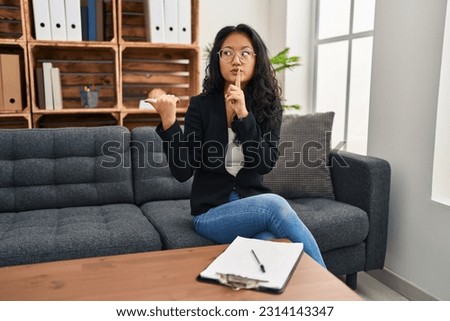 Young asian woman at consultation office asking to be quiet with finger on lips pointing with hand to the side. silence and secret concept. 