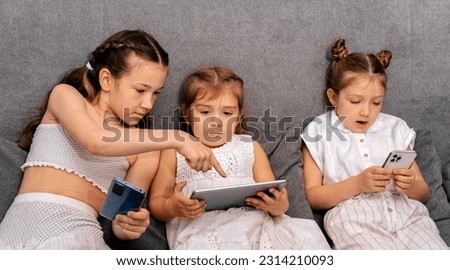 Cute little sisters sitting on sofa using modern gadgets to watch videos and play games.