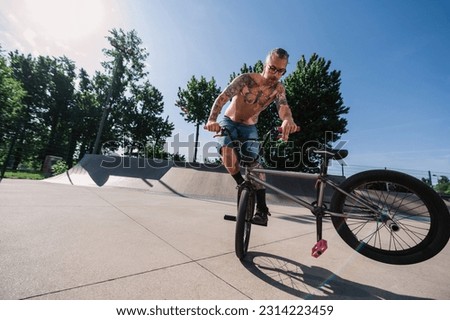 A shirtless tattooed mature man is performing tricks and stunts in a skate park with his bmx. A mature urban freestyle bike rider is practicing tricks and stunts on his bike in a skate park. Royalty-Free Stock Photo #2314223459