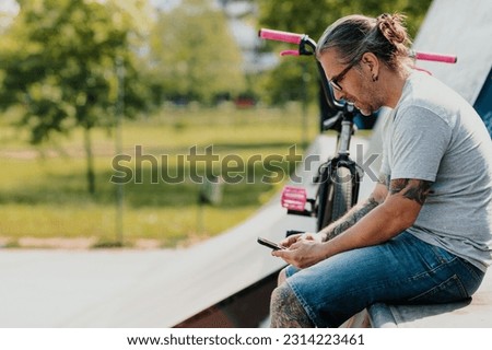 A middle-aged tattooed bmx bike rider is sitting in a skate park and typing messages on his mobile phone. Profile view of an older freestyle bike rider in a skate park using his phone.