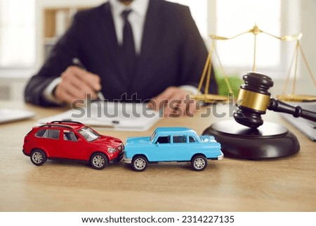 Close up of two toy model cars on background of judge who is conducting lawsuit in car accident. Toy cars representing accident stand on table next to judge's gavel the scales of justice. Royalty-Free Stock Photo #2314227135
