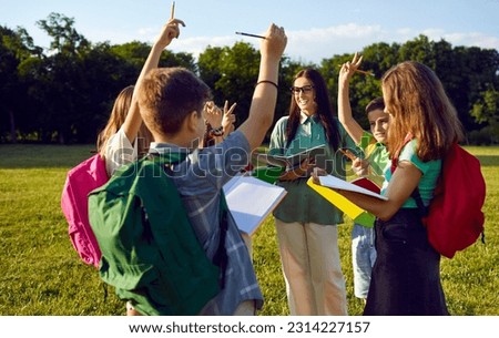 Happy school children having biology class outside. Group of classmates standing on green field, holding books and assignments, learning about nature, and raising hands to answer teacher's question Royalty-Free Stock Photo #2314227157