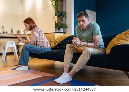 Unhappy angry husband sits on couch and feels heartache due wife obsession with smartphone, feels ignored and craves attention. Wife ignor husband. Jealous man afraid of betrayal. Toxic relationship Royalty-Free Stock Photo #2314244337