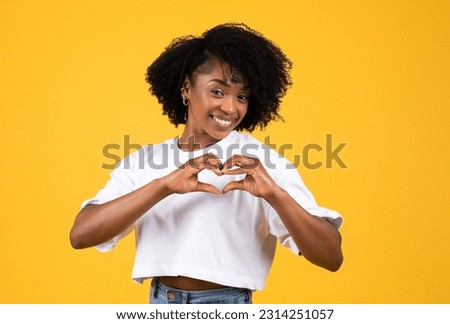 Positive millennial african american curly lady in white t-shirt make heart gesture with hands, isolated on yellow studio background. Lifestyle, student love, sign romantic, gratitude, ad and offer