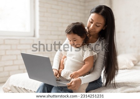 Baby And Gadgets. Japanese Mommy And Little Child Daughter Using Laptop, Browsing Internet For Online Fun Together Sitting In Cozy Bedroom Indoor. Mom And Toddler Watching Cartoons On Computer