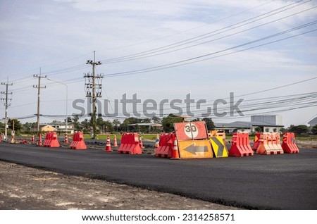 A view of the surface of a new black asphalt road with signs and orange barricades placed in the middle of a road being renovated near a power pole in rural Thailand during the day.