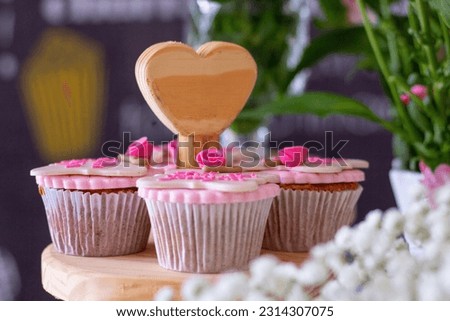 image with defocused green and white details, with cupcakes in focus with white and pink decor, on a wooden stand with a heart detail.
