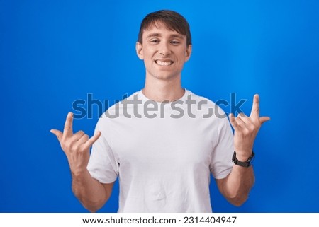 Caucasian blond man standing over blue background shouting with crazy expression doing rock symbol with hands up. music star. heavy music concept. 