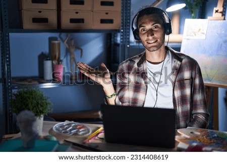 Young hispanic man sitting at art studio with laptop late at night smiling cheerful presenting and pointing with palm of hand looking at the camera. 