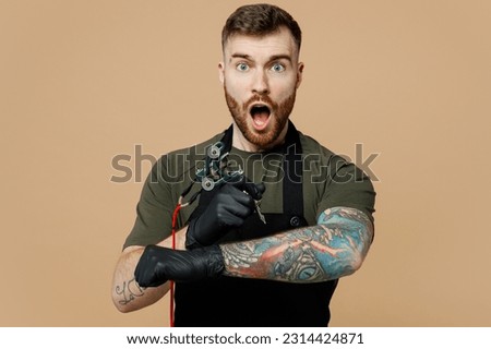 Surprised happy fun tattooer master artist tattooed man wear green t-shirt apron hold machine black ink in jar, equipment for making tattoo art on body for himself isolated on plain beige background
