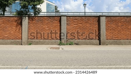 Surrounding brick wall with concrete columns with cloudy sky, street lamp and modern building on behind. Urban road in front. Background for copy space. Royalty-Free Stock Photo #2314426489