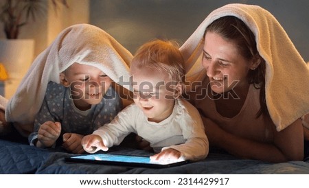 Happy smiling kids with mother lying under blanket and using tablet computer. Family having time together, parenting, happy childhood and entertainment