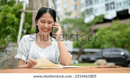A happy Asian woman in casual clothes is looking down at information in her book and talking on the phone with someone while sitting at a table outdoors.