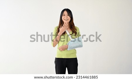 An amazed and surprised young Asian woman with a broken arm in an arm sling is smiling and covering her mouth while standing against an isolated white background.