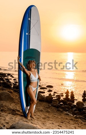 Graceful young woman wearing white bikini with a tattoo poses standing with a sup board on the beach. Beautiful sunset on background